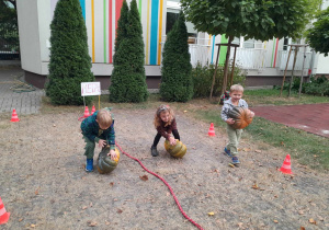 Na zdjęciu widać trójkę dzieci: dwóch chłopców i dziewczynkę. Dzieci turlają dynie po wyznaczonym torze. Z tyłu widać białą ścianę z kolorowymi paskami, trzy tuje i drzewo.