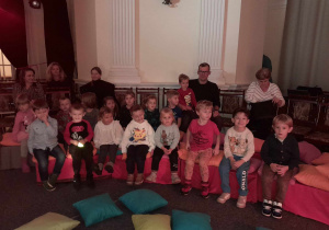 Na zdjęciu widać dzieci siedzące na czerwonych pufach. Za dziećmi siedzą dorośli. W tle widać ścianę z białymi filarami.