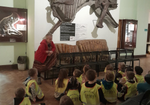 Na zdjęciu widać dzieci siedzące na podłodze. Dzieci są ubrane w jaskrawo żółte kamizelki. Obserwują szkielet dinozaura - eksponat w muzeum.