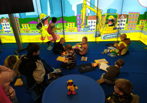 Zdjęcie dzieci siedzących na niebieskiej podłodze, oglądających prezenty otrzmane w trakcie zwiedzania i wizualizację przedstawiającą kolorowe rysunki metra i krecika w metrze.