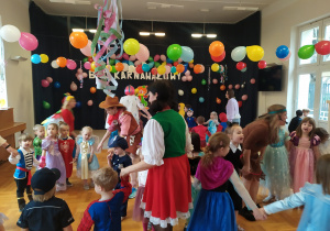 Na zdjęciu widać dzieci i nauczycielki w strojach karnawałowych, które tańczą ustawione w koła. W tle sala gimnastyczna przystrojona kolorowymi balonami. Na jednej ścianie na ciemnej kotarze widać girlandę z kolorowych balonów i napis "Bal karnawałowy"