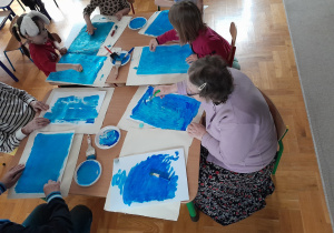 Przy stole siedzą dzieci i starsza kobieta. Na stole leżą białe kartki. Uczestnicy malują je niebieską farbą.