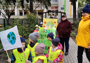 Na zdjęciu widać dzieci ubrane w kamizelki odblaskowe. Przedszkolaki ustawione w pary trzymają w rękach transparenty z hasłami ekologicznymi.