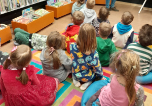 Na zdjęciu widać dzieci siedzące na kolorowym dywanie. W tle stoją półki z ustawionymi na nich książkami.