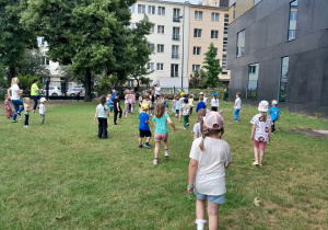 Na zdjęciu widać grupę dzieci biegnących na terenie pokrytym trawą. W tle widać drzewo i bloki mieszkalne.