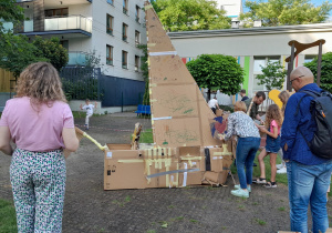 Na ziemi stoi żaglówka wykonana z kartonowych pudełek. wokół niej stoją rodzice i dzieci. w tle widać blok mieszkalny i budynek przedszkola.