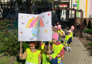 Na zdjęciu widać grupę dzieci ubranych w kamizelki odblaskowe. Dzieci stoją na chodniku, pierwsza para trzyma transparent z napisem "Czysta ziemia" oraz rysunkiem Ziemi trzymającej parasol. W tle widać przedszkole.