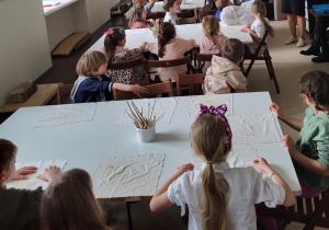 Dzieci siedzą przy białych stołach, przed każdym dzieckiem leży biały materiał. Na środku stołu stoi biały kubek z drewnianymi patyczkami.