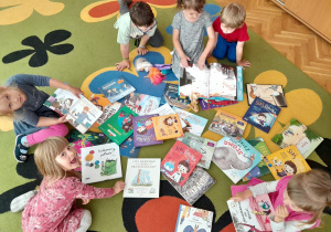 Na zdjęciu widać dzieci siedzące na kolorowym dywanie. Wokół nich rozłożone są różne książki, które dzieci przeglądają.