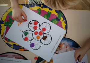Na zdjęciu widać stolik z kolorowymi podkładkami. Na pierwszym planie znajduje się biała kartka z kwiatkiem, który dzieci wyklejają kropkami z kolorowej plasteliny.